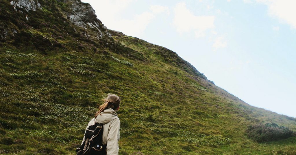 A woman wearing a dark backpack stares into the distance as she walks past a grassy hill.