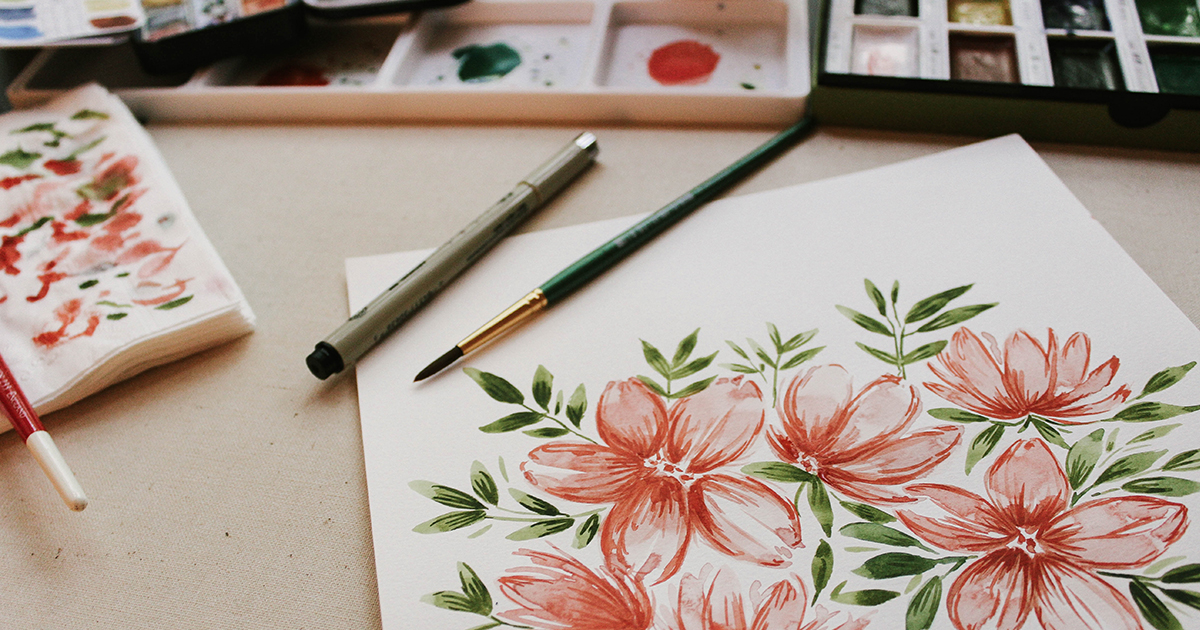 Una acuarela de flores rojas y hojas verdes sobre una mesa. El cuadro está rodeado de materiales de acuarela.