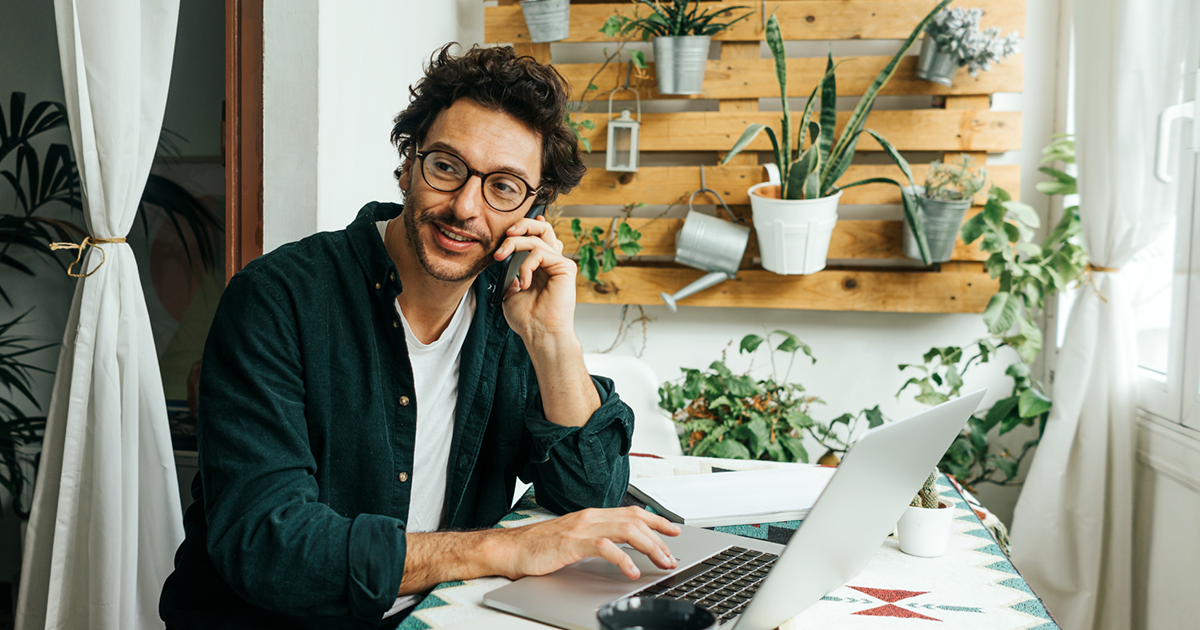 A man wearing glasses talks on the phone and uses a laptop while working from home.