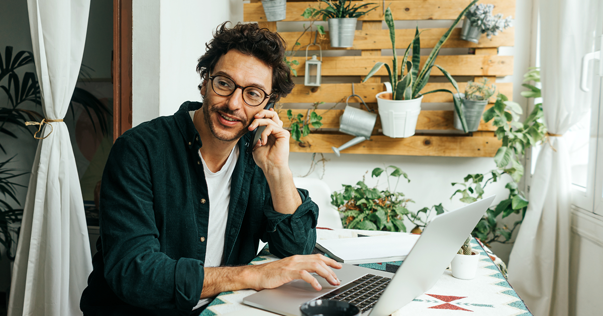 Un hombre con gafas habla por teléfono y utiliza un ordenador portátil mientras trabaja desde casa.