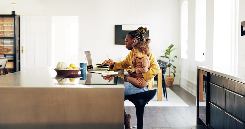 A black woman works on her laptop at a table while she holds her child on her lap.