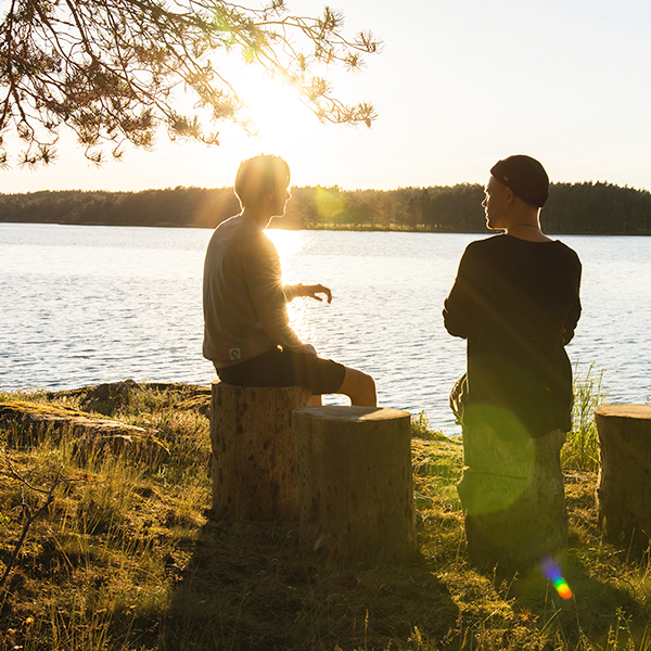Two men sit on stumps by a lake having a difficult conversation as the sun sets.