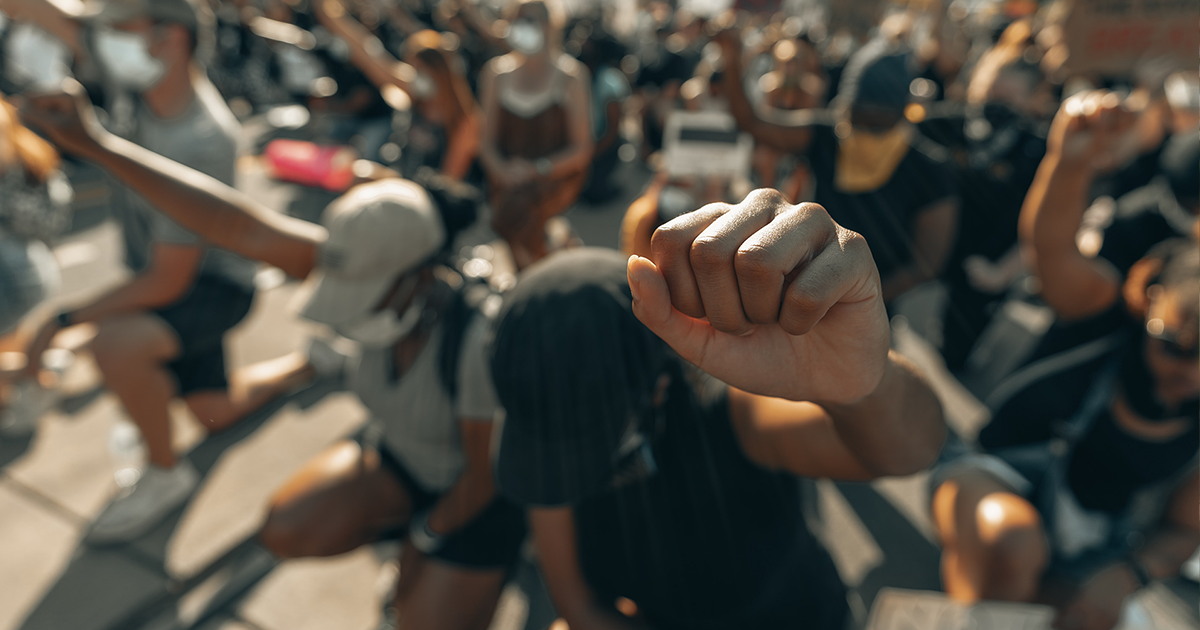 A Black individual wearing a baseball cap raises their fist while marching with a group.