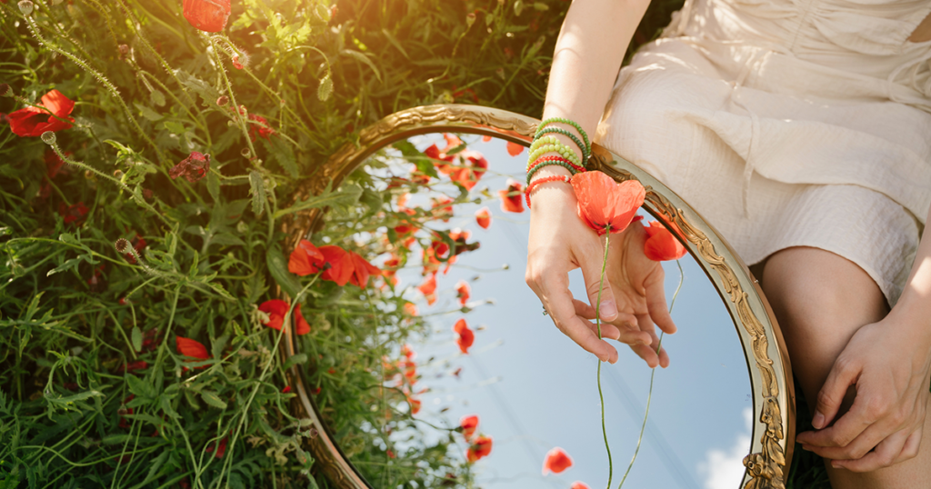 A woman in a cream dress sits in a field holding an orange flower over a vintage mirror.