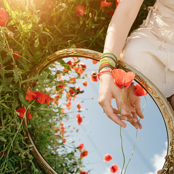 A woman in a cream dress sits in a field holding an orange flower over a vintage mirror.