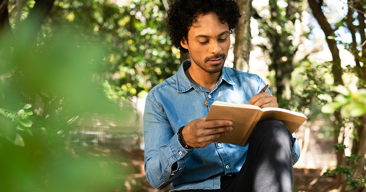 A man with curly black hair and a button down denim shirt sits outside among leafy green trees and writes in a journal.