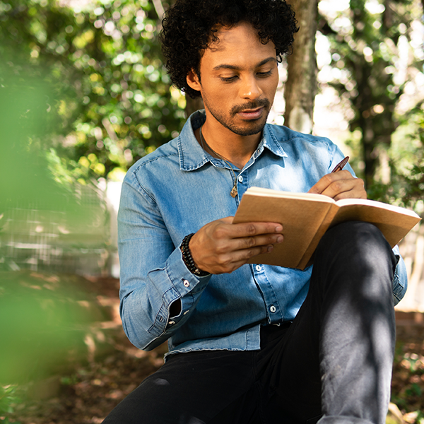 A man with curly black hair and a button down denim shirt sits outside among leafy green trees and writes in a journal.