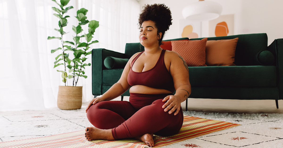 A Black woman wearing a maroon sports bra and leggings sits in her living room on a mat with her legs crossed, her eyes closed, and her hands on her knees.