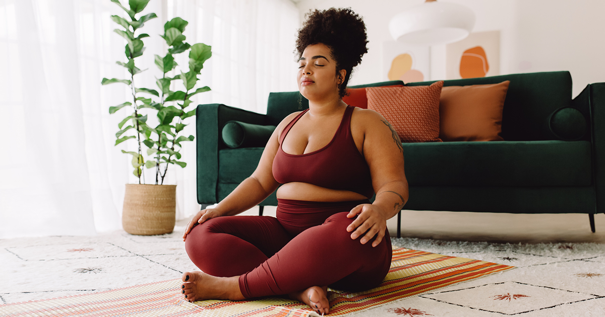 A woman wearing a maroon sports bra and leggings sits in her living room on a mat with her legs crossed, her eyes closed, and her hands on her knees.