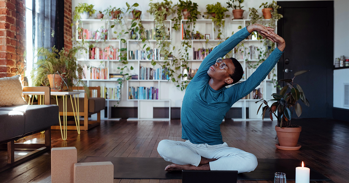 Un hombre se sienta en el suelo con las piernas cruzadas sobre una esterilla de yoga. Tiene los brazos extendidos por encima de la cabeza, con las manos juntas, mientras se estira hacia la izquierda.