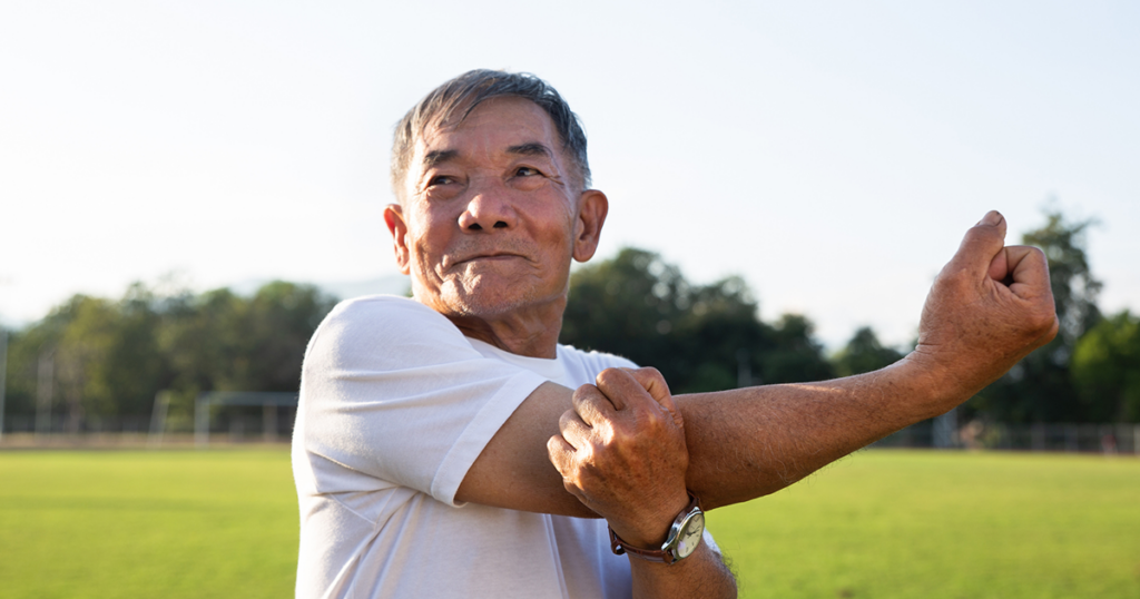 Un hombre con camiseta blanca está de pie en un parque y estira el hombro utilizando la mano izquierda para sostener el brazo derecho sobre el pecho.