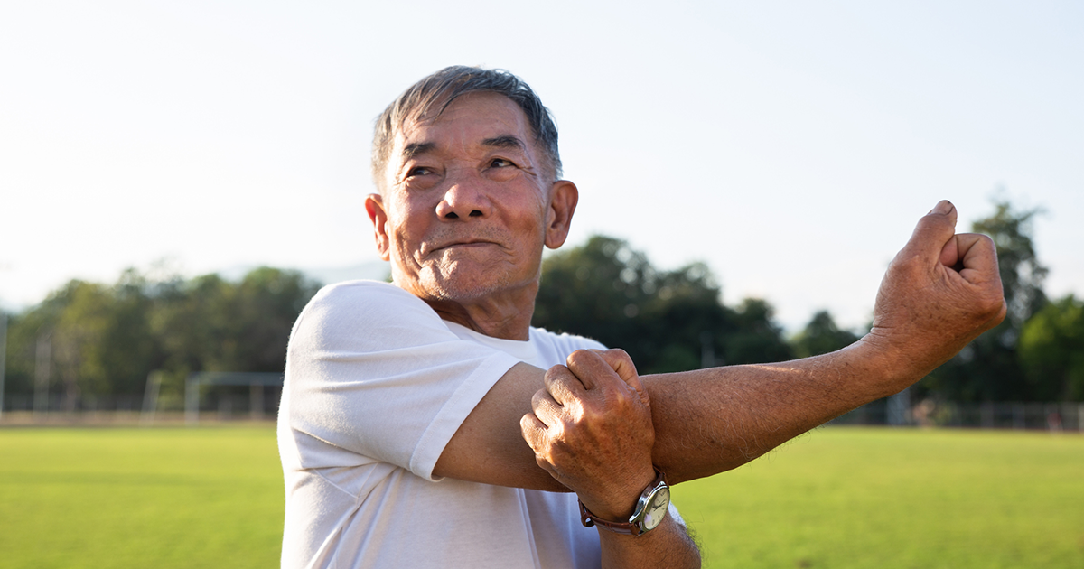 A man in a white t-shirt stands in a park and stretches his shoulder by using his left hand to hold his right arm across his chest.