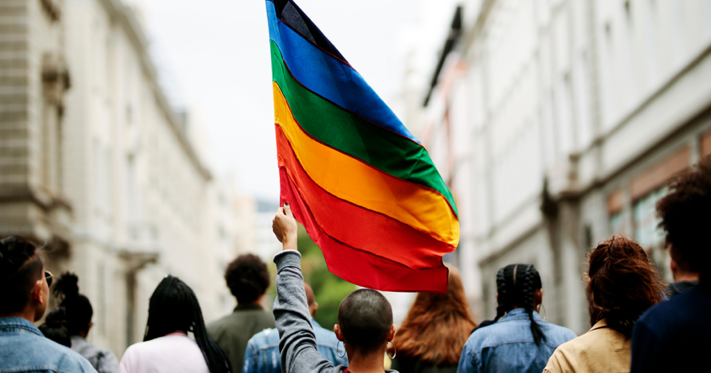 Un individuo camina entre una multitud sosteniendo una bandera arco iris sobre su cabeza.