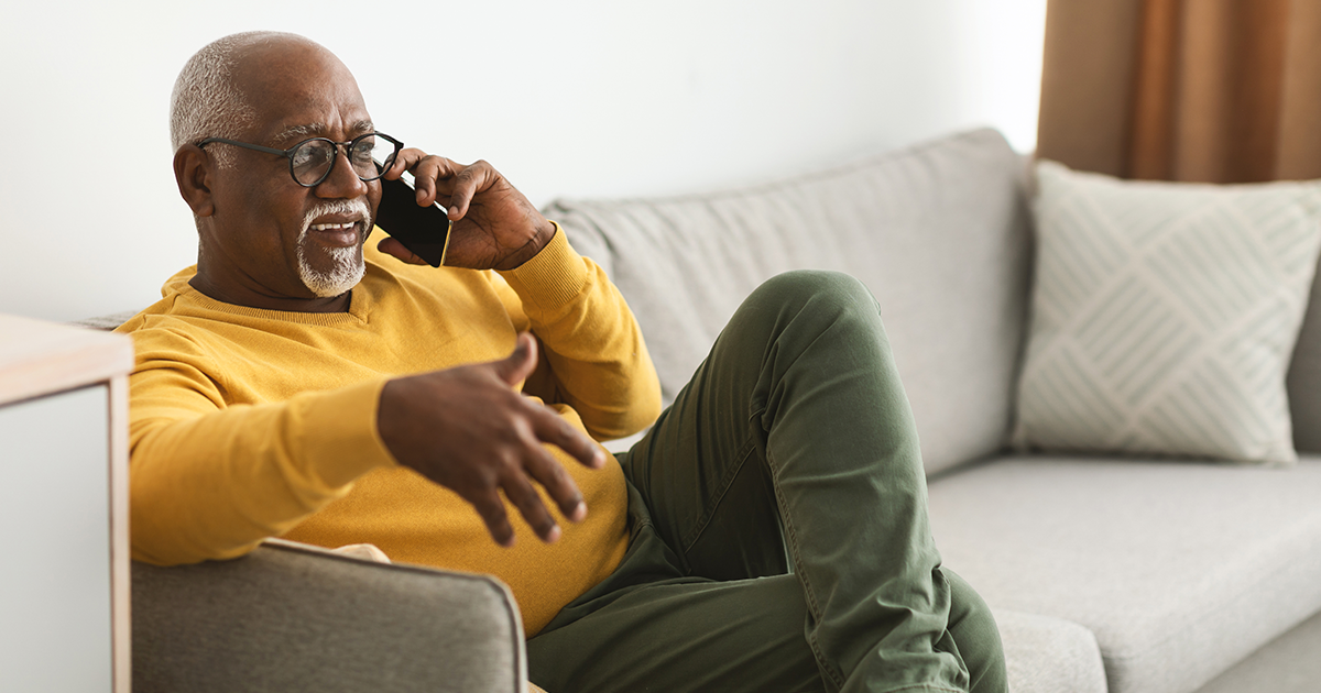 A man sitting on a couch talks on a cell phone while gesturing with his free hand.