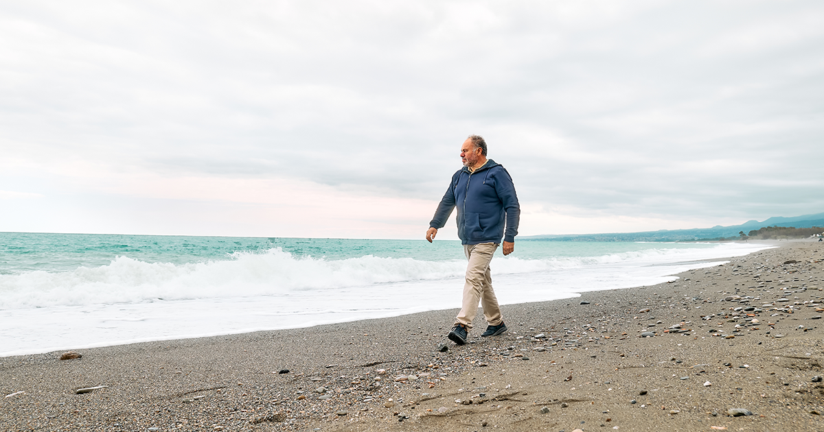 A man walks along a beach on a cloud day with waves lapping at the shore.