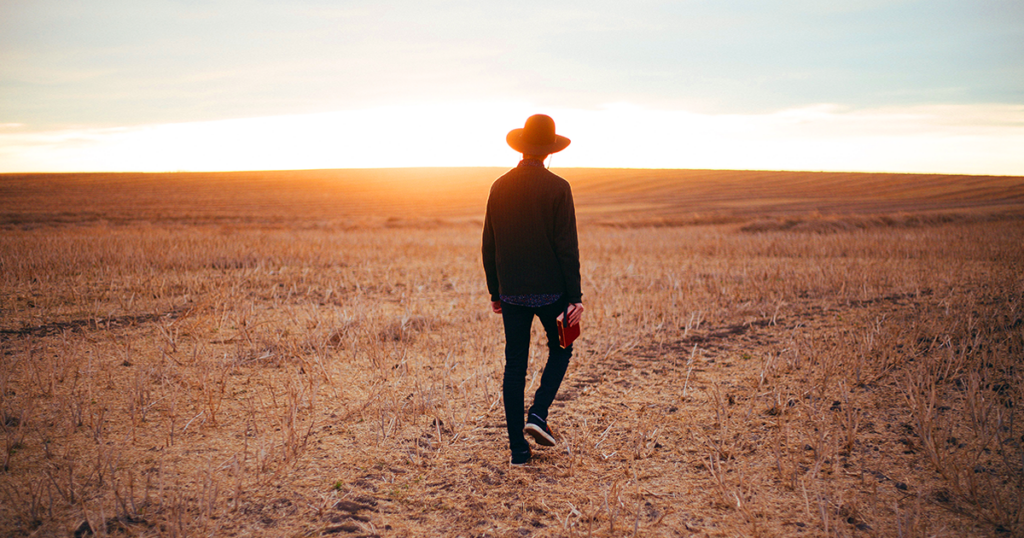 Un hombre con sombrero de ala ancha camina por un campo con un libro en la mano derecha.