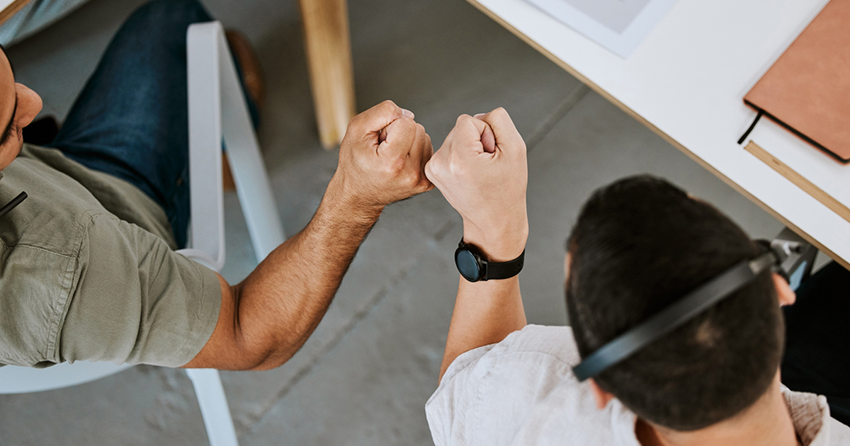 Two men sitting at a desk fist bump.