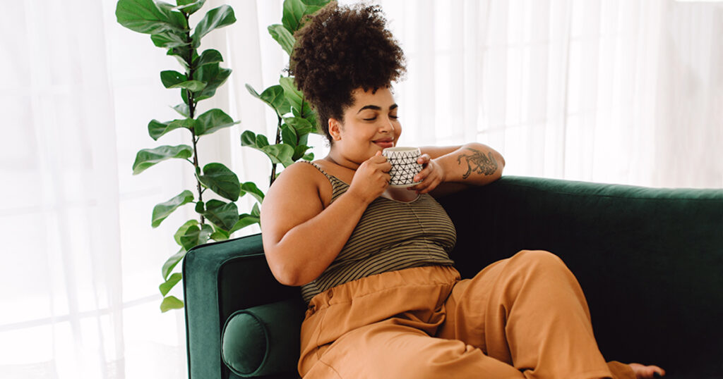A woman sits on a green couch smiling slightly with her eyes closed and both hands holding a mug close to her mouth.