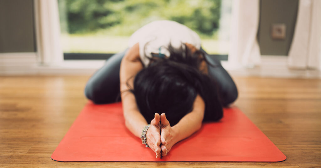 Una mujer se tumba en postura infantil sobre una esterilla de yoga roja con los brazos extendidos y las palmas juntas.