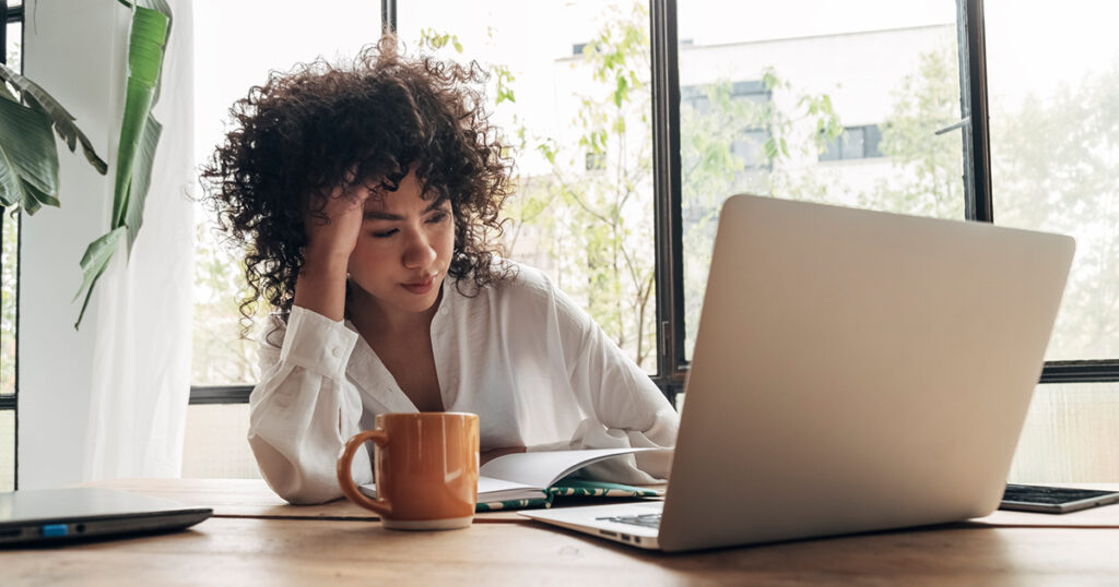 A woman sits at a table in front of a notebook and a laptop computer looking distraught.