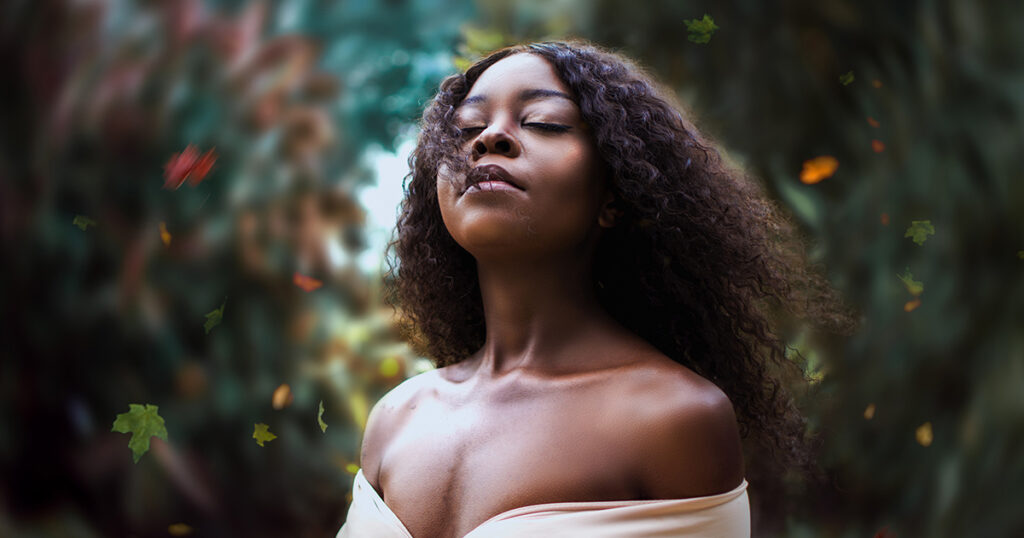 A Black woman stands in front of greenery with her chin lifted and her eyes closed.