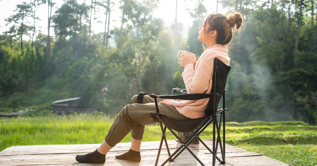 A woman sits in a chair outside holding a mug with her eyes closed.