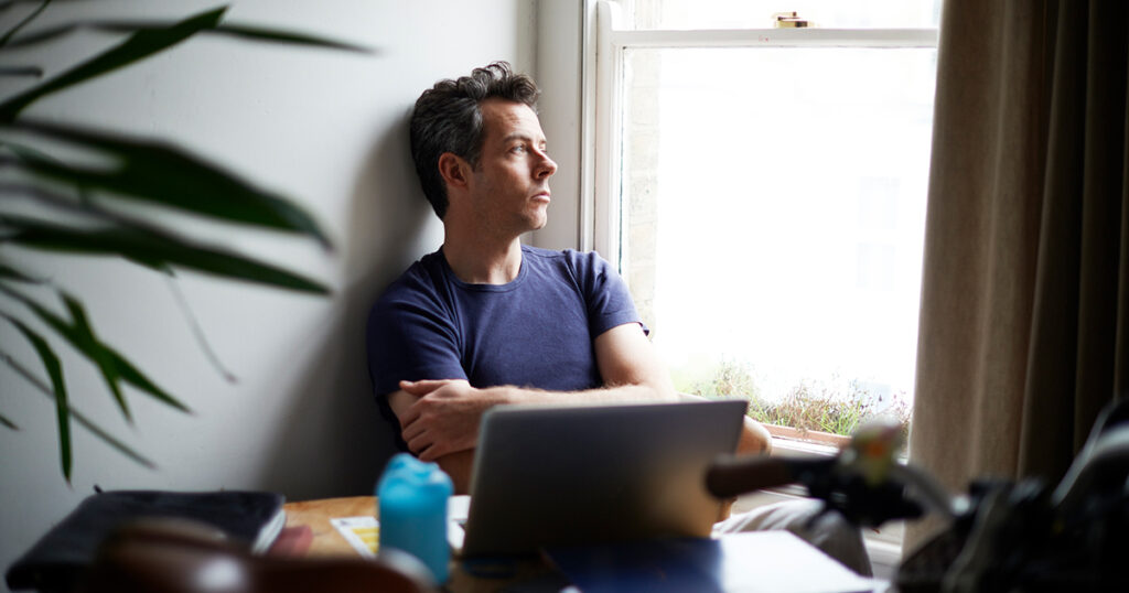A man sitting in front of an open laptop looks out a window with his arms crossed.