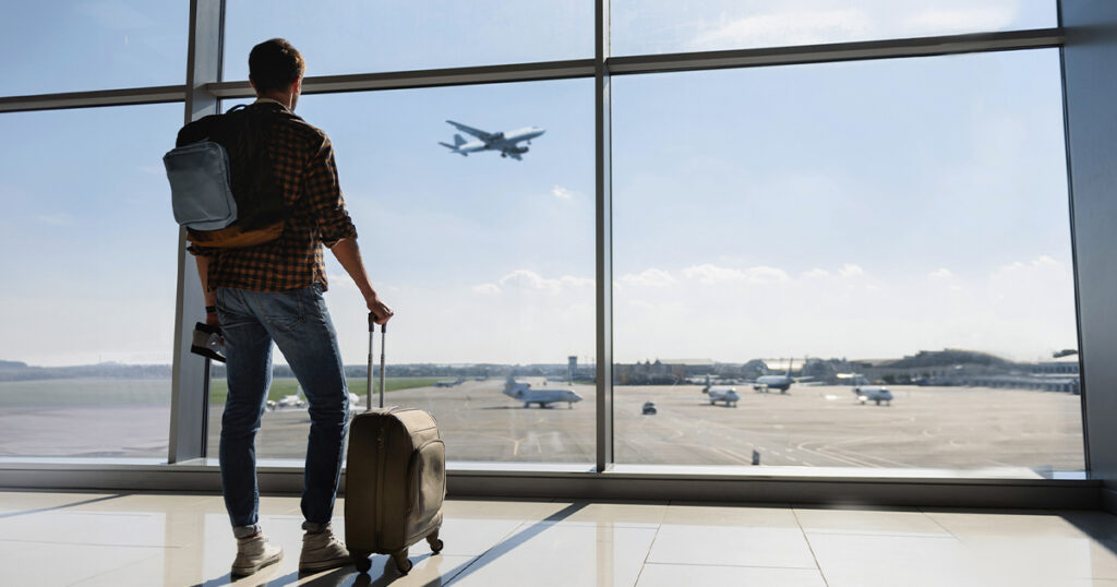 A man with luggage stands in front of a window in an airport watching a plane take off.