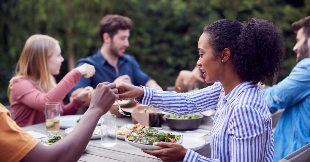 A woman eating outdoors with friends holds a salad and takes a fork from the hand of someone out of frame.