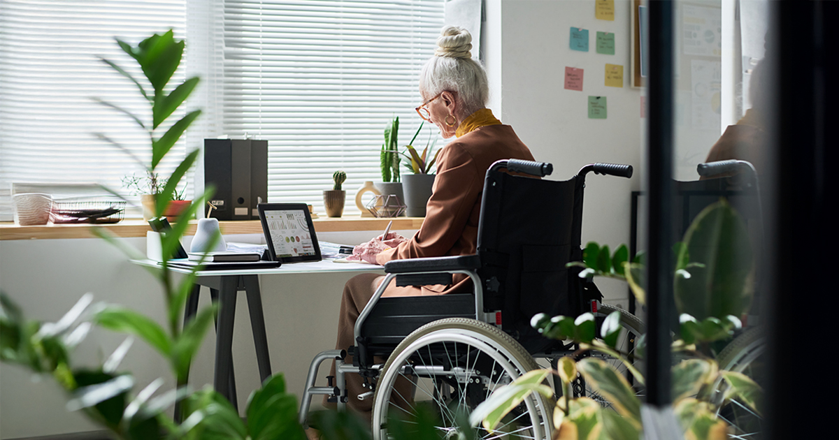 An older woman sits in a wheelchair, drawing at a desk.
