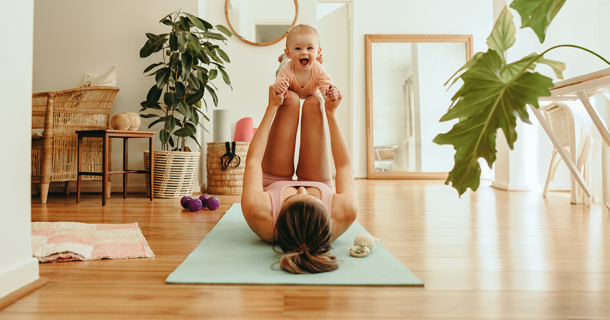 A mom lays on her back on a yoga mat with her knees bent in tabletop pose and her baby resting on her shins.