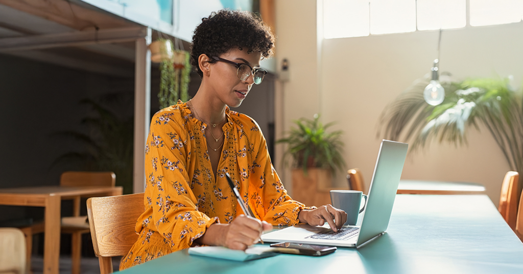 A woman sitting at a table works on her laptop while handwriting notes.