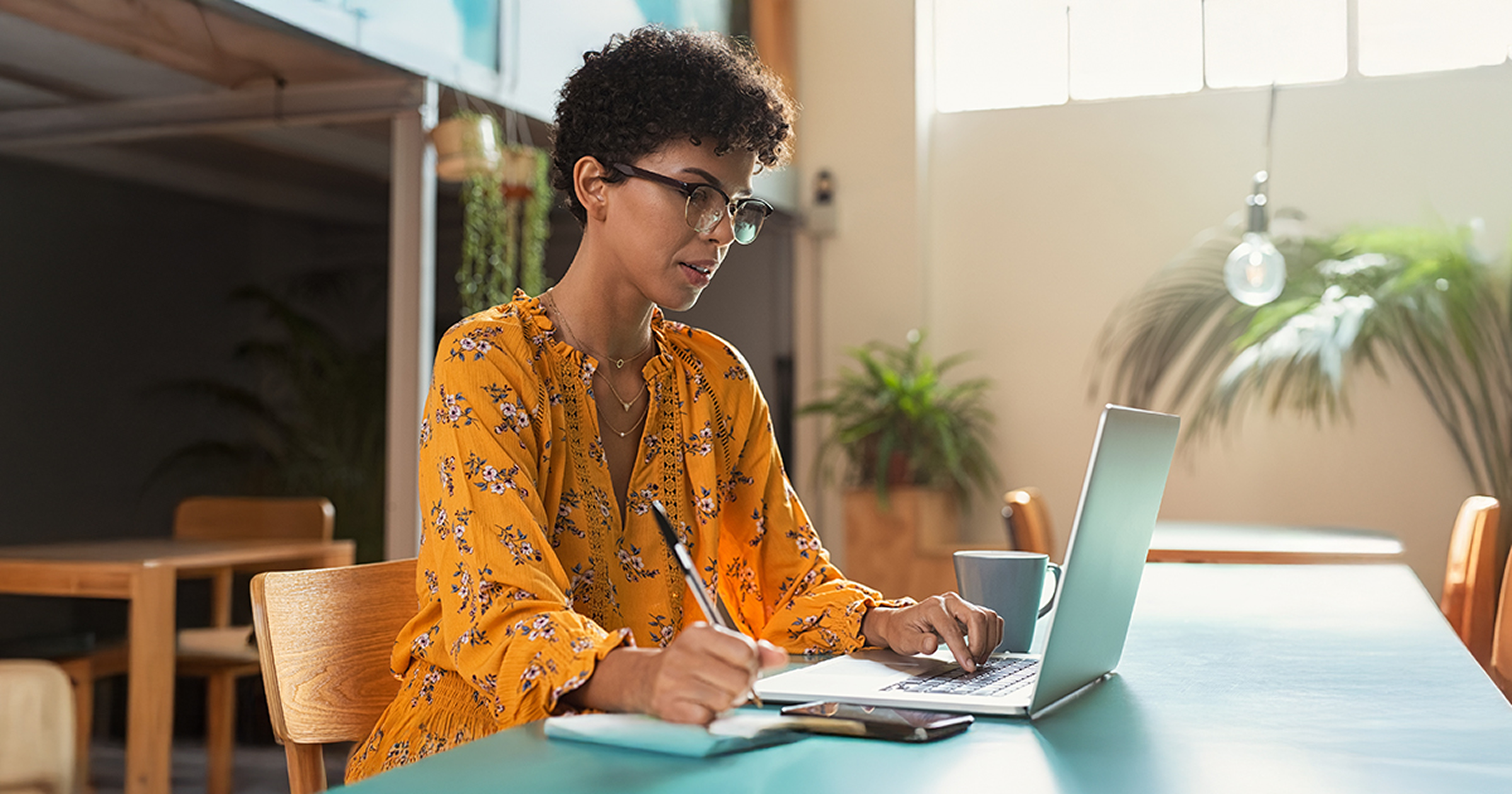 A woman sitting at a table works on her laptop while handwriting notes.