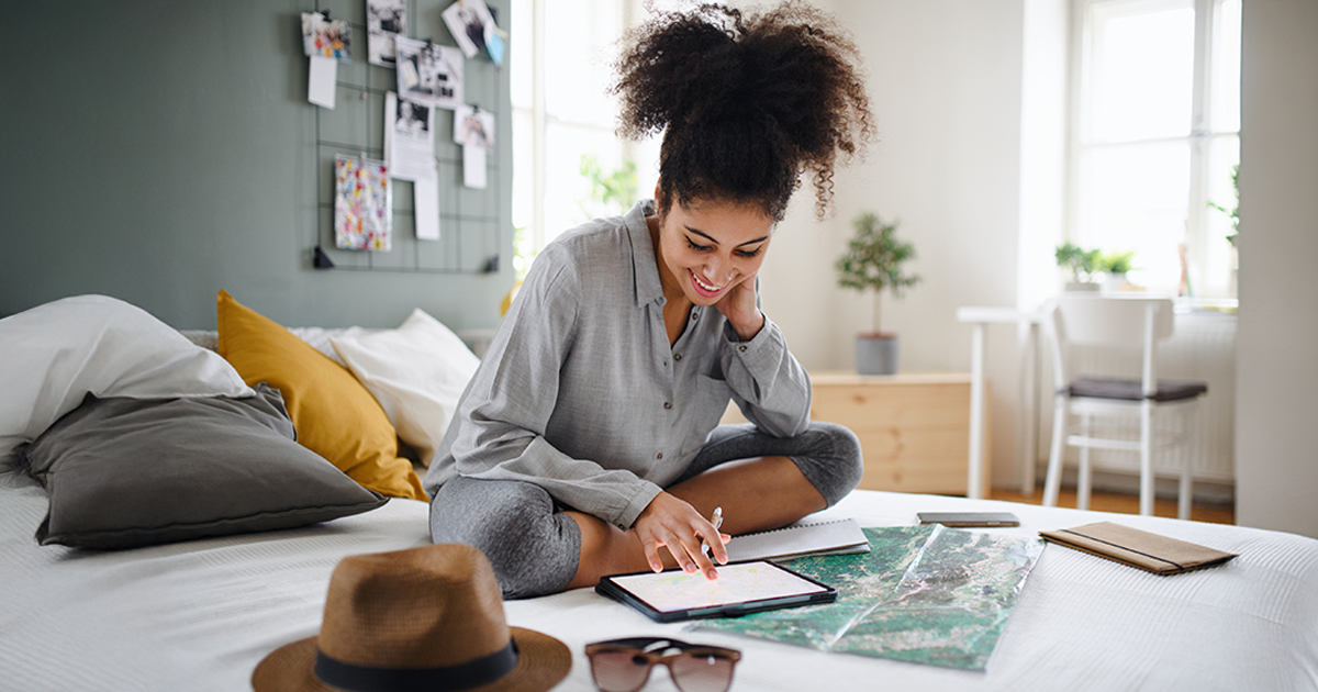A woman sits on a bed surrounded by a map, e-tablet, and travel accessories.