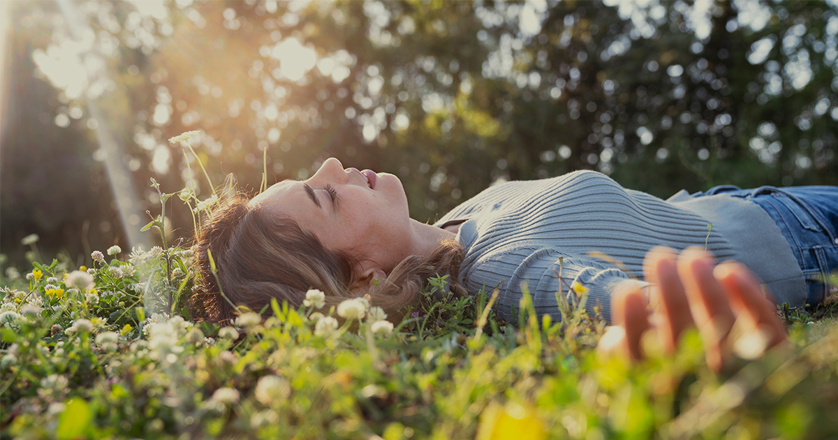 A woman in a blue sweater lays on her back in a sunny field.