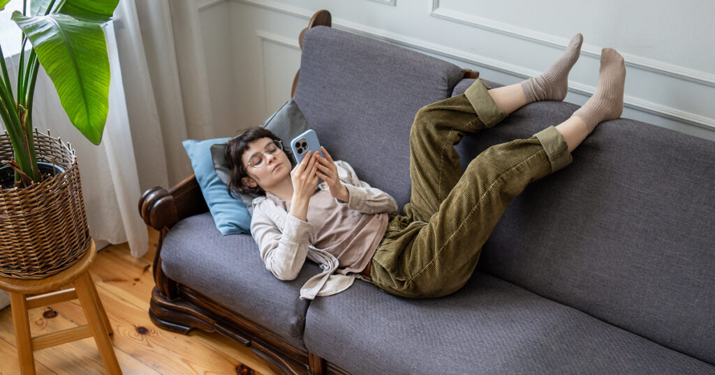 A woman looks at her phone while laying on the couch with her feet up against the back cushions.
