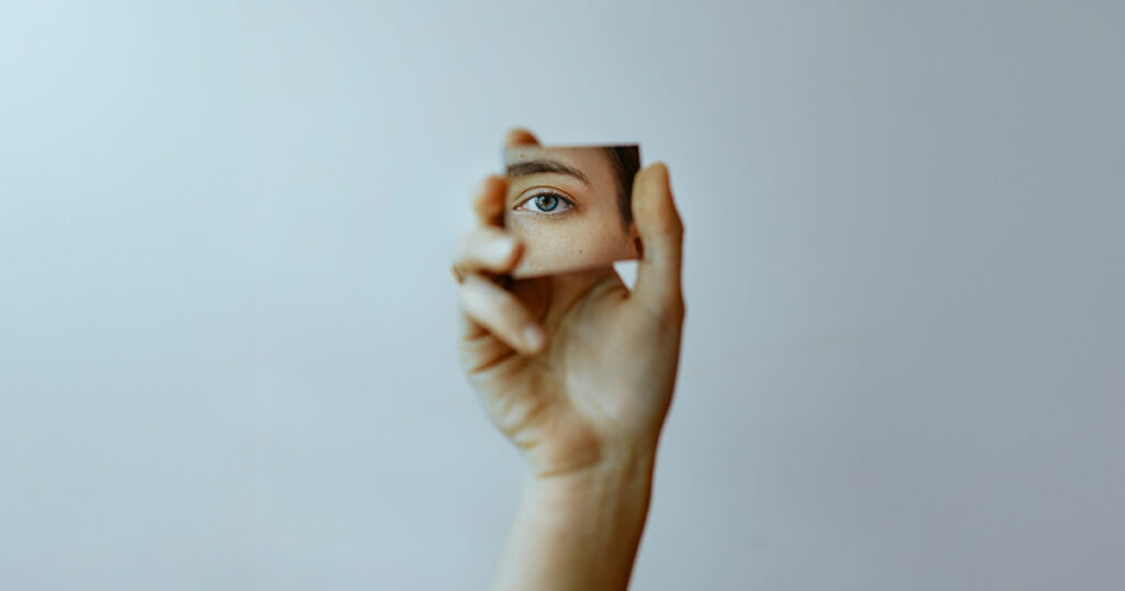 A person holds up a small square mirror that shows one eye.