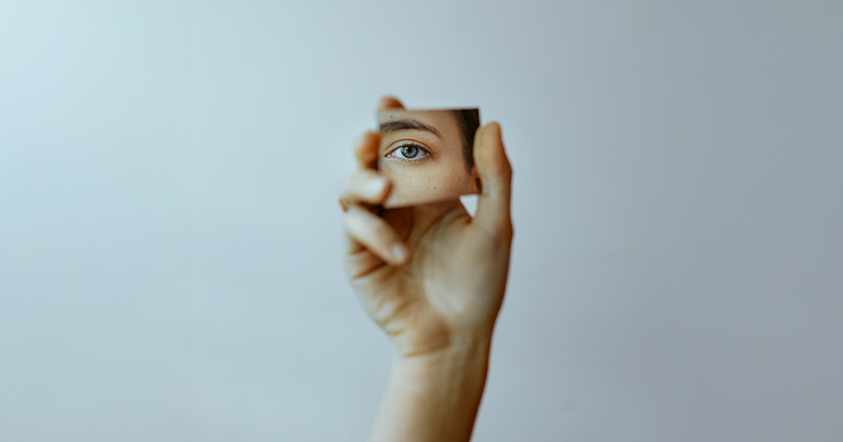 A person holds up a small square mirror that shows one eye.