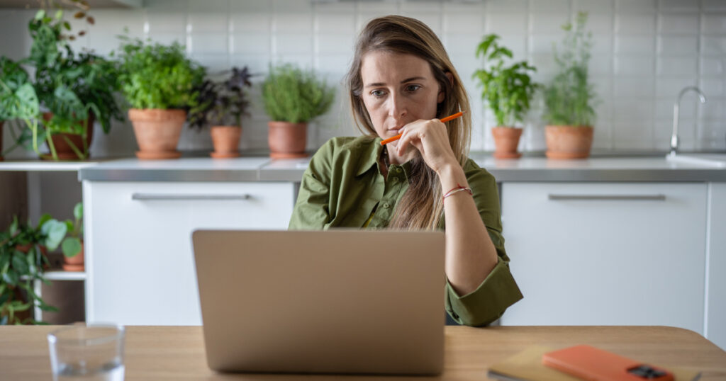 A woman stares at a laptop screen while holding an orange pen.
