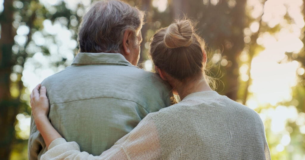 A woman puts her arm around an older man while outside.